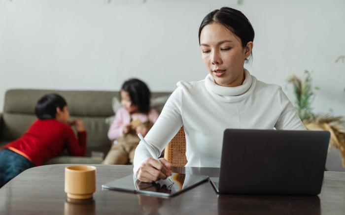 Woman in white turtleneck sitting at her kitchen table writing something down with laptop in front of her. Her two children play int the background.