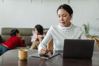 Woman in white turtleneck sitting at her kitchen table writing something down with laptop in front of her. Her two children play int the background.